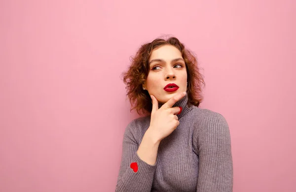 Funny curly girl with thoughtful face stands on pink background and looks away at Copy space and thinks. Closeup portrait of a thoughtful hipster girl in a gray sweater and with makeup. — Stockfoto