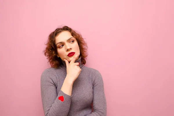 Pensive girl in gray sweater and curly red hair isolated on pink background, looking away at blank space and thinking. Portrait of a thinking cute lady looking at copy space. Isolated. — Stockfoto