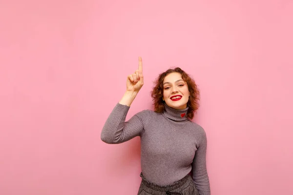 Menina adolescente sorridente com cabelo encaracolado e em uma camisola cinza fica em um fundo rosa e mostra seu dedo indicador para cima em um ponto em branco, olha para a câmera com um rosto feliz. Menina alegre recomenda — Fotografia de Stock