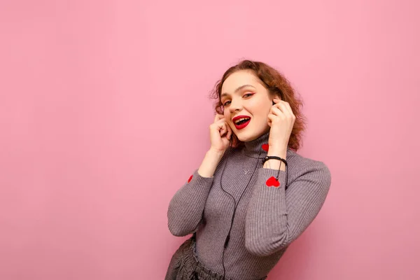 Retrato de senhora bonita feliz em camisola cinza e cabelos cacheados ouvindo música em fones de ouvido sobre fundo pastel rosa, olhando para a câmera e sorrindo. — Fotografia de Stock