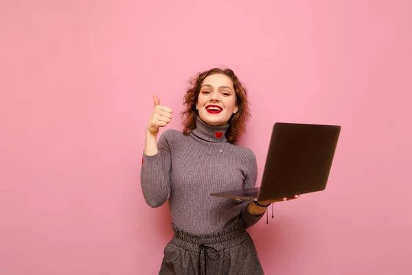 Весела кучерява дівчина в сірому повсякденному одязі стоїть з ноутбуком в руці на рожевому тлі, дивиться в камеру з посмішкою на обличчі і показує її великий палець вгору. Щаслива леді з ноутбуком ізольовано — стокове фото