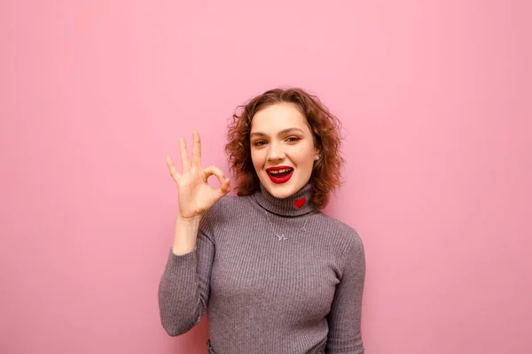 Happy lady in casual kleding en met krullend rood haar geïsoleerd op een roze pastel achtergrond, toont Ok gebaar, kijkt in de camera en glimlacht. Geïsoleerd. Kopieerruimte — Stockfoto