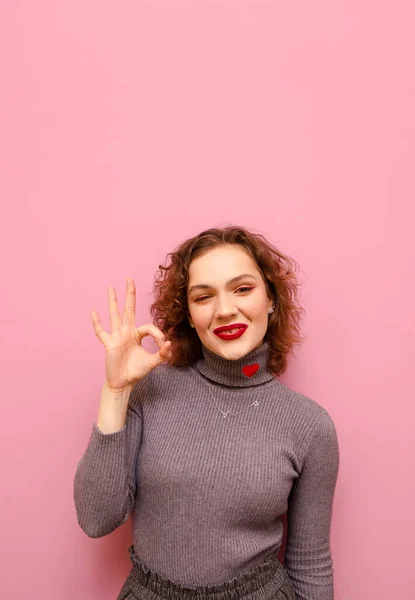 Jovem senhora com cabelo encaracolado e camisola cinza mostra um gesto de mão OK e olha para a câmera com um sorriso, retrato vertical em um fundo rosada.Menina estreita os olhos e mostra o gesto TODO BOM — Fotografia de Stock