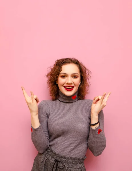 Smiling curly redhead girl isolated on pink background, shows heavy metal gesture and looks into camera with happy face, wears gray casual clothes with hearts. Vertical photo. — ストック写真