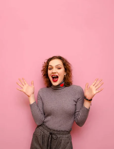 Retrato de estúdio de senhora alegre em roupas casuais e com cabelos vermelhos encaracolados em fundo rosa pastel, olha para a câmera com rosto surpreso e se alegra. Menina alegre emocionalmente alegra-se. Vertical — Fotografia de Stock