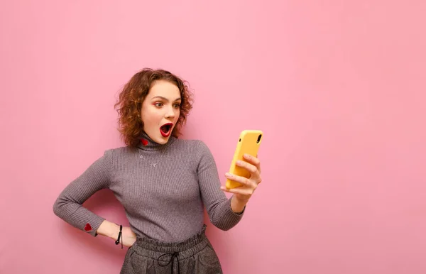 Retrato de estudio de una linda chica emocional usando un teléfono inteligente con la cara impactada sobre un fondo rosa pastel. Sorprendida dama atractiva en ropa casual con estilo utiliza un teléfono inteligente. Aislado . — Foto de Stock