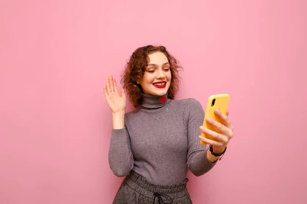 Мила усміхнена дівчина з кучерявим волоссям робить відеодзвінок на смартфон і показує жест привітань співрозмовника, ізольовані на рожевому пастельному тлі. Концепція відеозв'язку. Копіювати простір — стокове фото