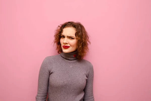 Cute girl with curly hair and casual clothes looks suspiciously into the camera on a pink background. Funny lady with red hair and gray sweater looks into the camera and suspects isolated on pink — ストック写真