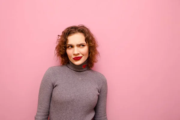 Menina bonito com um olhar suspeito e cabelo encaracolado vermelho isolado em um fundo pastel rosa, olha para o espaço de cópia com um rosto atencioso. Isolados . — Fotografia de Stock