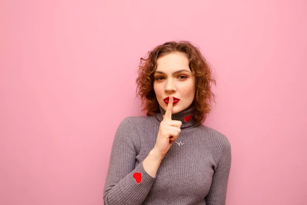 Menina adolescente bonito com cabelo encaracolado vermelho colocar o dedo nos lábios, mostra um sinal de silêncio, e olha para a câmera com um rosto sério em um fundo pastel rosa. Senhora treme o gesto de Shsh — Fotografia de Stock