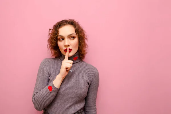 Cute girl with curly red hair put her finger to her lips, shows a gesture of silence, and looks away at copy space with a serious face on a pink pastel background. Lady shows the gesture of Shsh — ストック写真