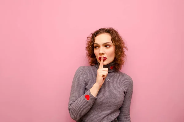 Cute girl with curly red hair has put her finger on her lips, shows a gesture of silence, and looks into the camera with a serious face on a pink pastel background. Lady shows the gesture of Shsh — ストック写真