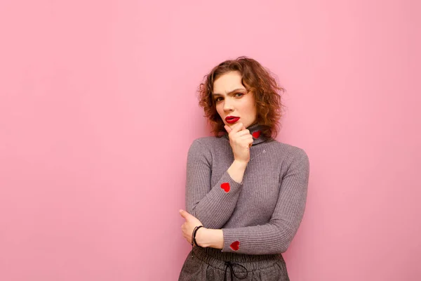 Menina adolescente cuidadosa com cabelo encaracolado vermelho e em uma camisola cinza fica em um fundo rosa e olha atentamente para a câmera e pensa. Pensive jovem senhora isolada no fundo pastel . — Fotografia de Stock