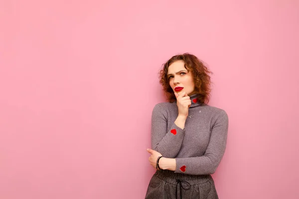 Menina adolescente cuidadosa com cabelo encaracolado vermelho e em uma camisola cinza fica em um fundo rosa e olha para o espaço de cópia, vestindo roupas casuais. Pensive jovem senhora isolada no fundo pastel — Fotografia de Stock