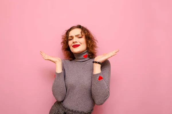 Funny curly hipster girl in gray sweater spreads her arms and looks confusedly at camera on pink background. Cheerful lady isolated on a pink background, with a smile on her face looking in camera — Stockfoto