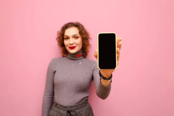 Smartphone with black blank screen in hands of beautiful girl. Positive girl in gray clothes and makeup stands on a pink background with a smartphone in her hand, looks in camera and smiles.Copy space — Stock Photo, Image