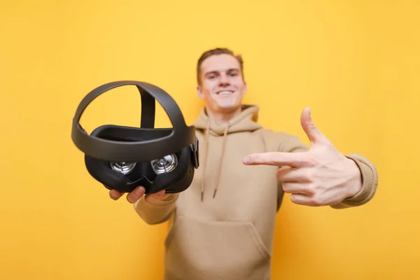 El joven sostiene un casco de realidad virtual en su mano, le señala con el dedo y sonríe. El tipo feliz muestra su dedo en el casco VR en su mano sobre un fondo amarillo.. Enfoque en el gadget VR . —  Fotos de Stock
