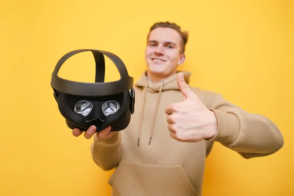 Happy young man holding a VR helmet in his hand, showing thumbs up, looking into the camera against a yellow background. Guy recommends a VR gadget in his hand. Isolated. Focus on glasses in the hands — 스톡 사진