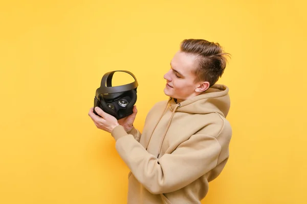 Portrait of a young man in a pink sweater stands with a VR helmet for playing on a yellow background and looking a gadget with smile.Guy with VR glasses isolated on yellow background. Virtual reality — 스톡 사진