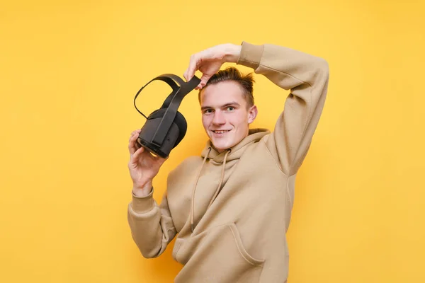 Retrato de cara feliz no fundo amarelo, de pé com capacete VR nas mãos, olhando para a câmera e sorrindo. Jovem positivo experimentando capacete realidade virtual, isolado. VR jogos . — Fotografia de Stock