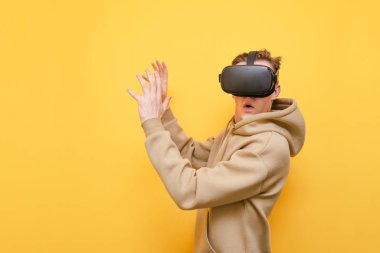 Surprised young man in VR clothes wearing VR glasses and with shocked face looks into camera on yellow background. Emotional guy is using Virtual Reality Helmet for the first time. Isolated.