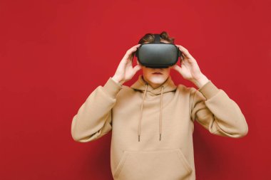 Portrait of a guy in casual clothes and with a VR helmet on his head on a red background with a serious face. Young man uses VR helmet, isolated. Copy space