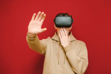 Portrait of a shocked guy in casual clothes and VR helmet on a red background, covers his mouth with surprise and shows his hand to the camera. Surprised guy in virtual reality helmet touches hand.