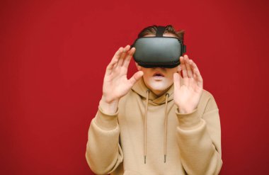Portrait of a shocked guy in a VR helmet on a red background, covers his face with his hand and looks in camera with a surprised face, wearing casual clothes. Emotional teen uses VR eyeglasses