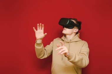 Portrait of a young man in casual clothes testing a VR helmet on a red background, looking away and touching his hands. Guy in the virtual reality helmet is isolated on a yellow background.
