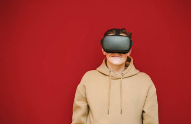 Closeup portrait of funny guy wearing virtual reality helmet on head, looking into camera and laughing, wearing beige hoodie isolated on red background. Cheerful guy in VR glasses playing games.