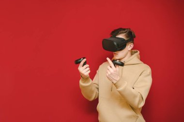 Teen gamer in a VR helmet stands on a red background with controls in his hands and plays virtual reality games, looking away with a focused face. Guy playing VR games with glasses, isolated.