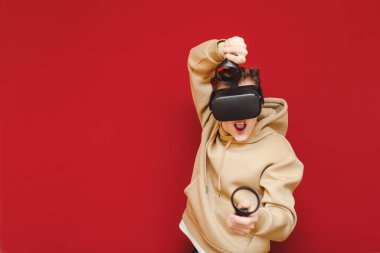 Cheerful guy stands with controllers in his hands and VR helmet on his head against a red background and plays video games with a funny face. Young gamer playing VR games with helmet and smiling.