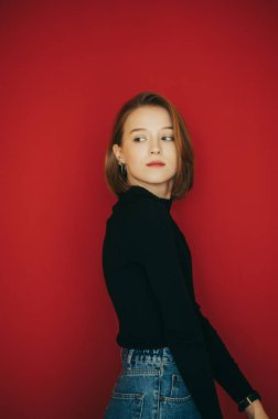 Stüdyoda kırmızı arka planda izole edilmiş siyah kazaklı güzel genç bir manken, başka tarafa bakıyor ve kameraya poz veriyor. Çekici bir kadının dikey portresi..