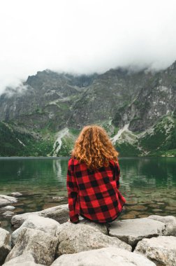Kırmızı gömlekli kadın dağlardaki güzel bir gölde taşların üzerinde oturur ve manzaraya bakar. Tatra Dağları 'ndaki Morskie Gölü' nün arka planında kızıl saçlı kıvırcık bir kızın arkası.