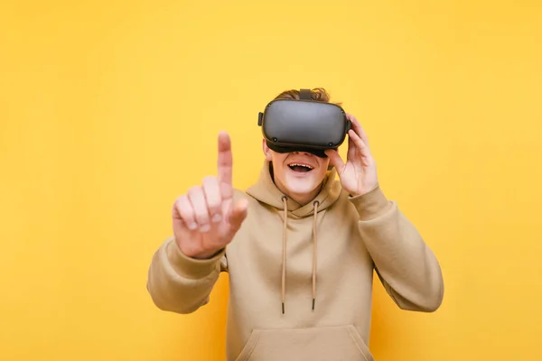 Joyeux jeune homme debout sur un fond jaune avec un casque VR sur la tête, souriant et touchant son doigt à la caméra. Le gars dans un casque de réalité virtuelle touche son doigt avec une image dans des lunettes — Photo