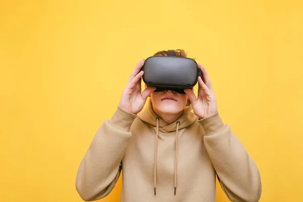 Un jeune homme en pull beige porte un casque de réalité virtuelle sur fond jaune. Portrait d'un gars en lunettes VR. Isolé. Concept de jeu VR . — Photo