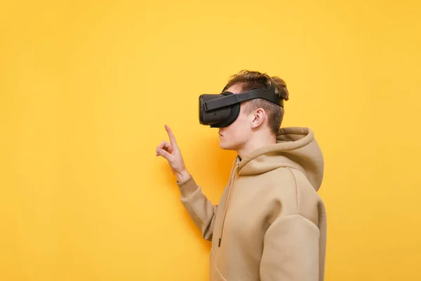 Calme jeune homme debout sur un fond jaune avec un casque VR sur la tête, touchant son doigt à côté d'un espace de copie. Le gars dans un casque de réalité virtuelle touche son doigt avec une image dans des lunettes — Photo