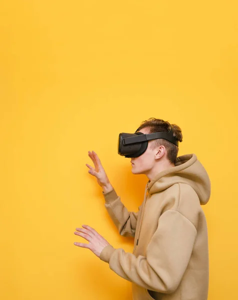 Photo verticale d'un jeune homme portant un casque VR sur fond jaune, regardant loin l'espace de copie et touchant ses mains. Homme dans un casque de réalité virtuelle isolé sur fond jaune . — Photo