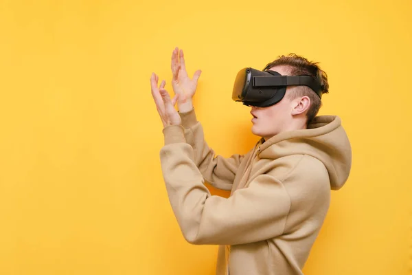 Portrait de gars surpris dans le casque VR se tient sur fond jaune et regarde ses mains. Jeune homme en vêtements décontractés utilise un casque de réalité virtuelle. Isolé . — Photo