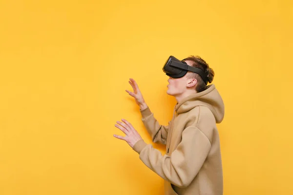 Jeune homme choqué dans le casque VR isolé sur fond jaune, regarde loin et touche les mains pour copier l'espace. Surprenant gars utilise un casque VR et est étonné de la réalité virtuelle, portant des vêtements décontractés — Photo