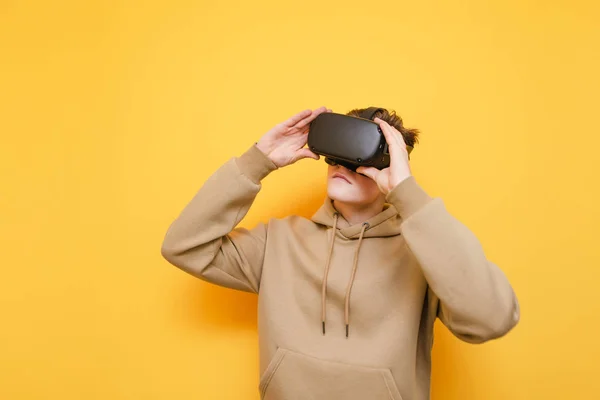 Jeune homme en sweat à capuche léger et casque VR se tient sur un fond jaune et regarder loin de l'espace de copie avec un visage sérieux.Jeune homme en réalité virtuelle casque isolé sur jaune portant des vêtements décontractés — Photo