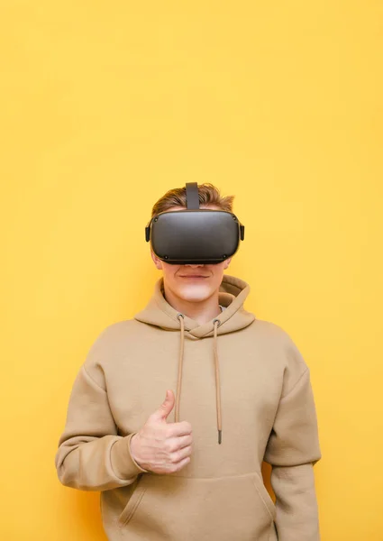 Adolescent positif gars en réalité virtuelle casque et vêtements décontractés isolés sur fond jaune, montrant pouces vers le haut et souriant.Jeune homme souhaitant utiliser la technologie VR, porte un casque. — Photo