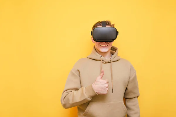 Jeune homme heureux en tenue décontractée et casque VR se tient sur un fond jaune, regarde dans la caméra avec un sourire et montre son pouce vers le haut, le gars aime la réalité virtuelle. Isolé. Espace de copie — Photo