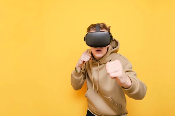 Jeune homme drôle en vêtements décontractés et un casque VR se tient dans un rack de boxe sur un fond jaune et regarde dans la caméra. Un gars concentré jouant avec un casque VR. Isolé . — Photo