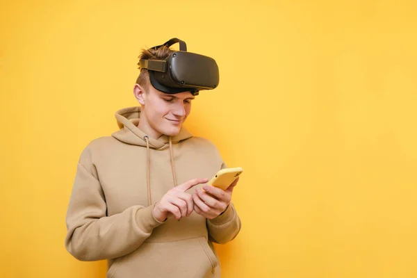 Cheerful guy in virtual reality helmet on his head isolated on yellow background with smartphone in his hands, looks into the screen with a smile on his face. Isolated.