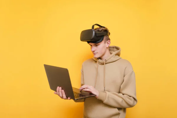 Skoncentrowany młody człowiek w luźnym ubraniu i kasku Vr na głowie stoi z laptopem w rękach na żółtym tle i korzysta z internetu.Guy w kasku wirtualnej rzeczywistości używa komputera. — Zdjęcie stockowe