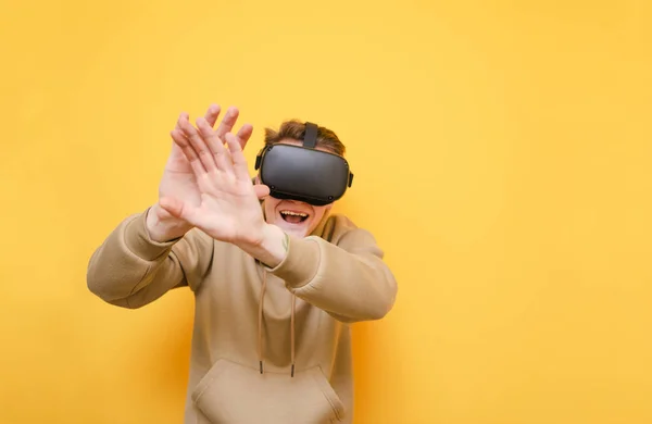 Jeune homme émotionnel sur fond jaune, portant un casque VR et des vêtements décontractés, souriant et couvrant son visage de ses mains. Joueur expressif joue à des jeux VR en réalité virtuelle casque, isolé . — Photo