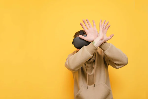 El joven asustado en casco VR cubre la cara con las manos del realismo de realidad virtual aislado sobre fondo amarillo. Tipo emocional en ropa casual jugando juegos de realidad virtual. Copiar espacio — Foto de Stock