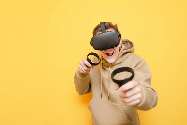 Cara alegre fica com controladores em suas mãos e capacete VR em sua cabeça contra um fundo amarelo e joga videogames com um rosto engraçado. Jovem jogador jogando jogos VR com capacete e sorrindo . — Fotografia de Stock