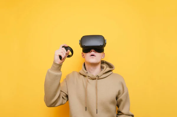 Portrait de jeune homme surpris dans le casque VR se tient sur fond jaune avec contrôleur à la main et lève les yeux dans l'étonnement. Guy gamer joue à des jeux vidéo sur casque VR. Isolé . — Photo